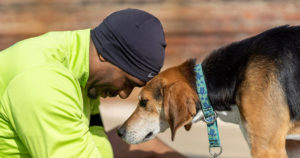 Man in running clothes sitting on the sidewalk, head lowered to touch foreheads with a tan and black dog