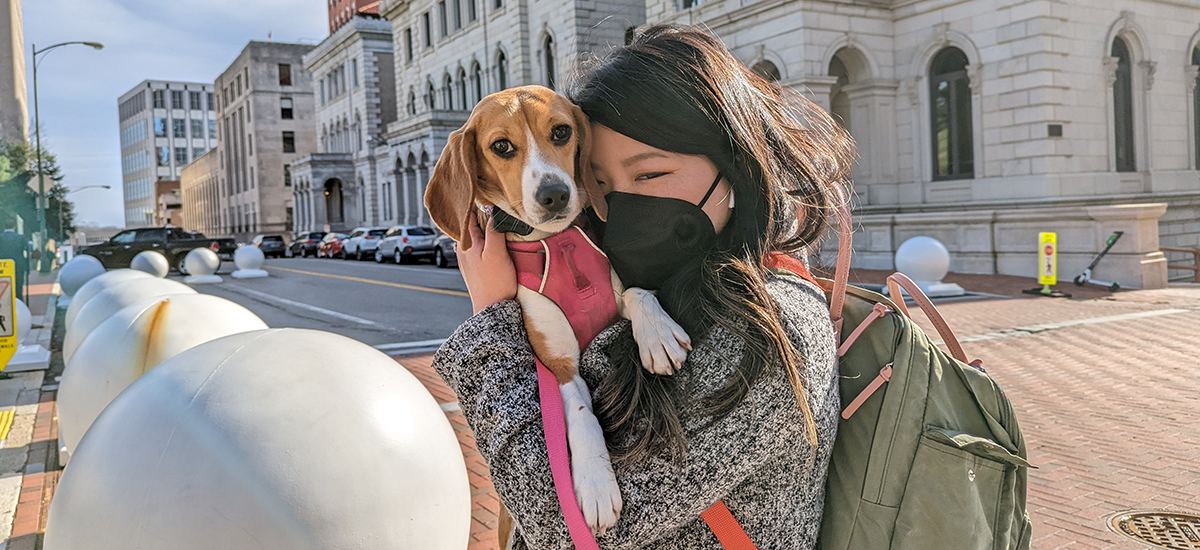 Virginia legislator stands on Bank Street outside the Virginia General Assembly holding a small beagle wearing a pink collar. The woman is wearing a black KN95 mask and presses her forehead to the dog's soft ear.