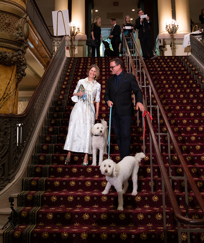 Two large, white, fluffy dogs stand on a wide, carpeted staircase accompanied by a man and woman in formal attire
