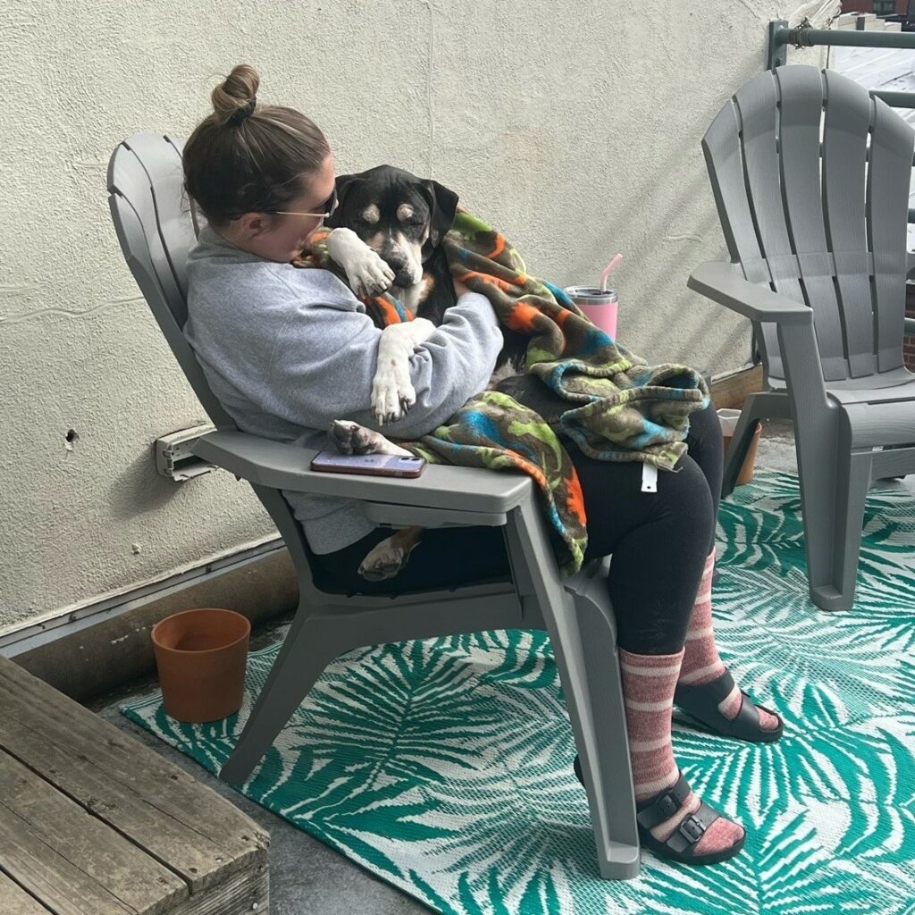 Woman sitting in a patio chair holding a medium-sized black and tan dog who is wrapped in a colorful blanket